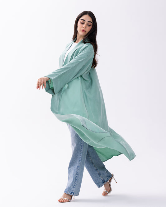 Woman wearing a light green long garment over a white top and blue jeans on a white background
